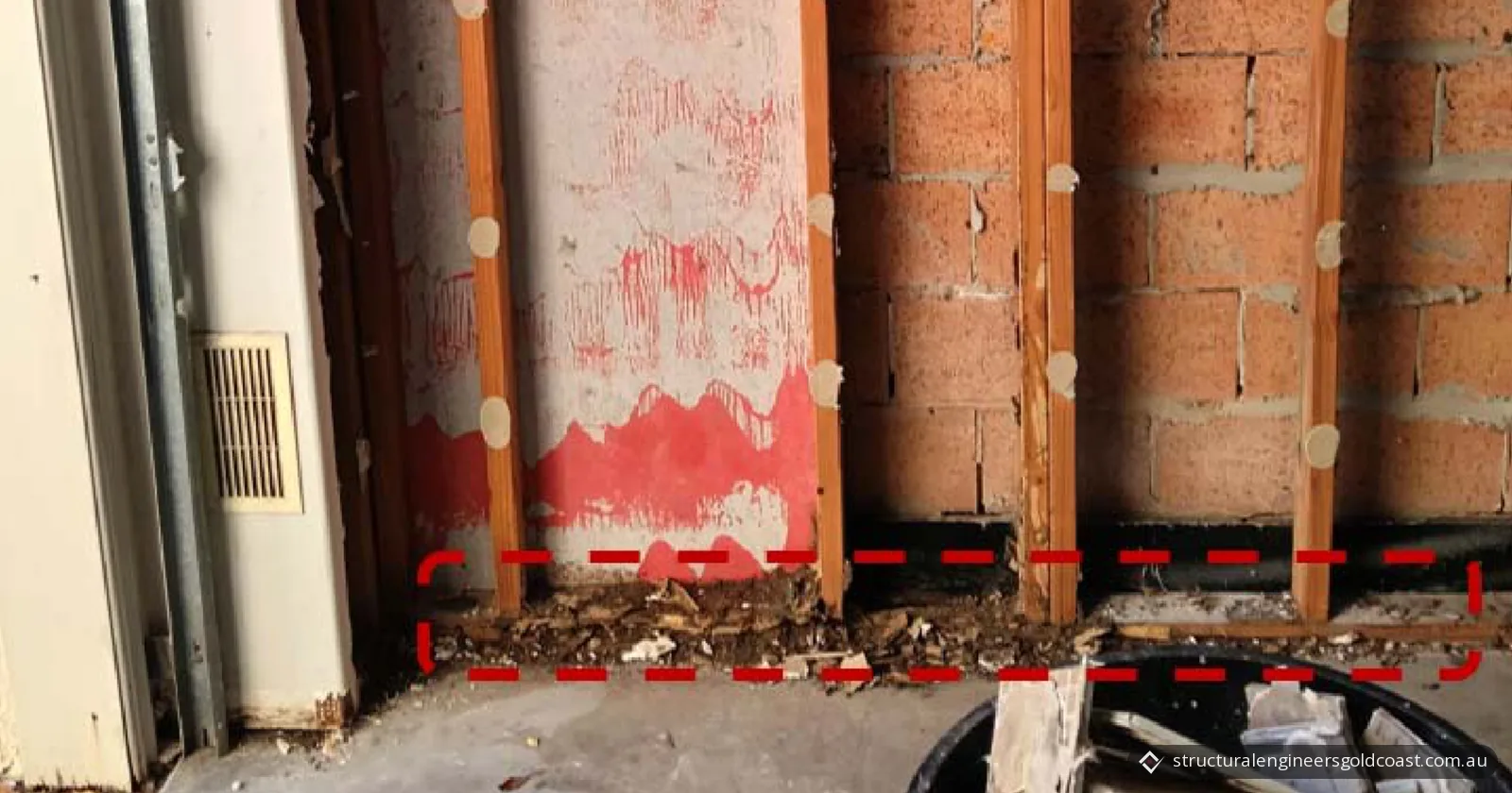 Base of a garage wall with the lining removed, showing timber studs against masonry and crumbled timber along the floor inside a red dashed box